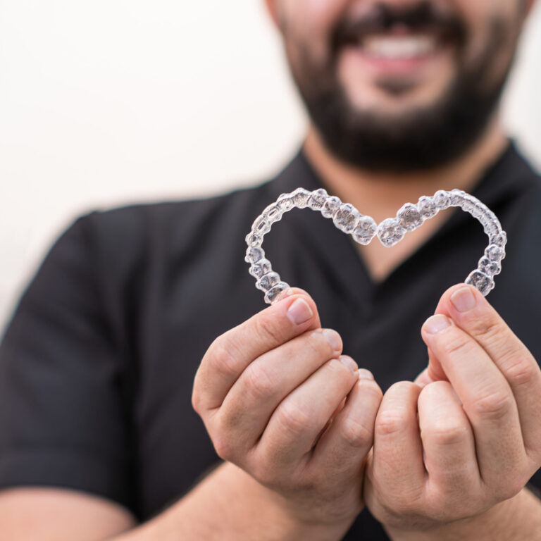 Smiling doctor holding heart made of clear aligners for professional teeth correction in stomatology clinic orthodontist showing love and care of patients closeup