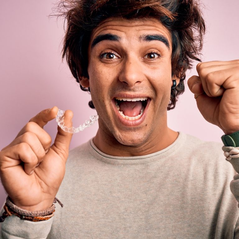 Young handsome man holding aligner standing over isolated pink background screaming proud and celebrating victory and success very excited, cheering emotion